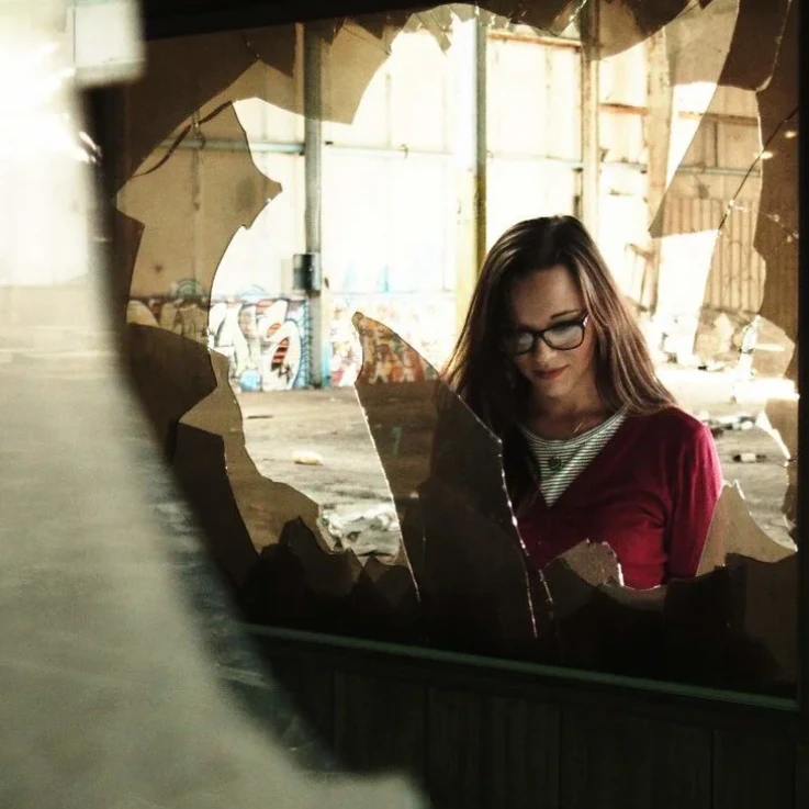 A woman reflected in broken glass inside an abandoned building.