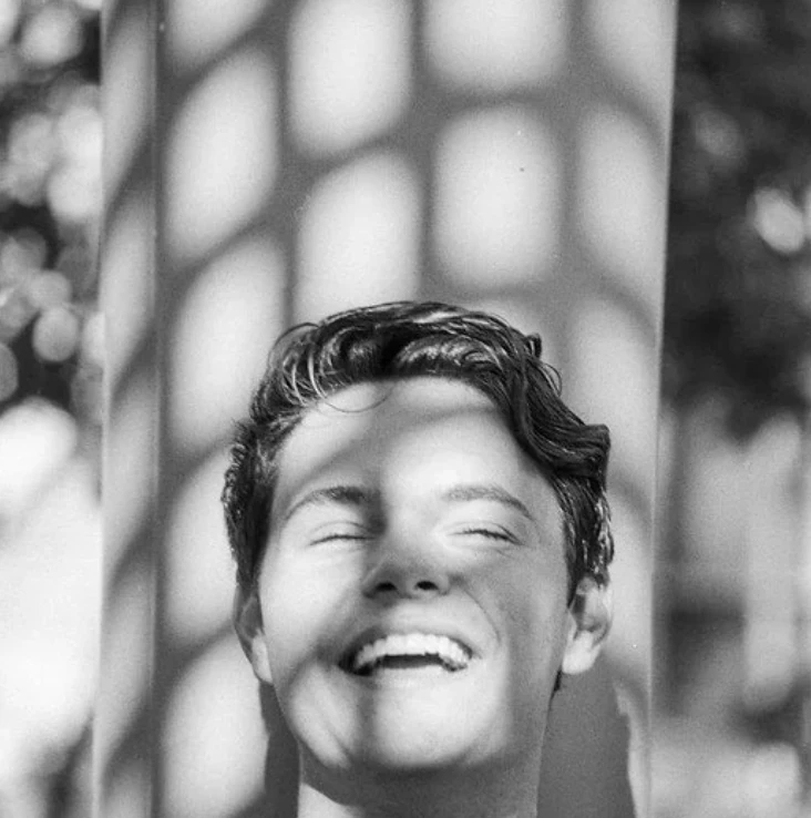 Black-and-white portrait of a person laughing in patterned sunlight.