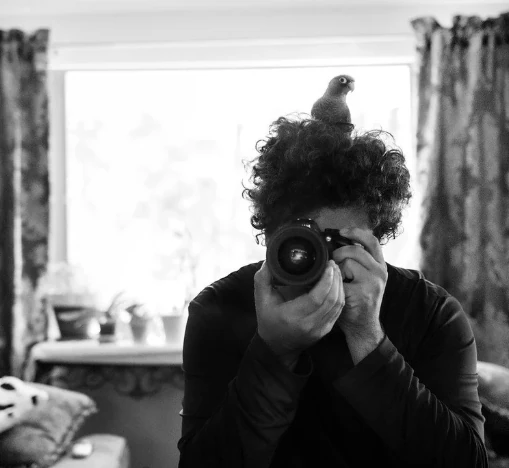 Black-and-white self-portrait of Trace Myers holding a camera, with a small bird perched on their hair.